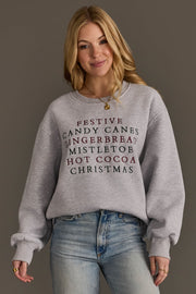 A woman wears a grey Christmas List Embroidered Sweatshirt featuring an oversized fit and embroidered text listing holiday favorites like festive, candy canes, gingerbread, mistletoe, hot cocoa, and christmas.