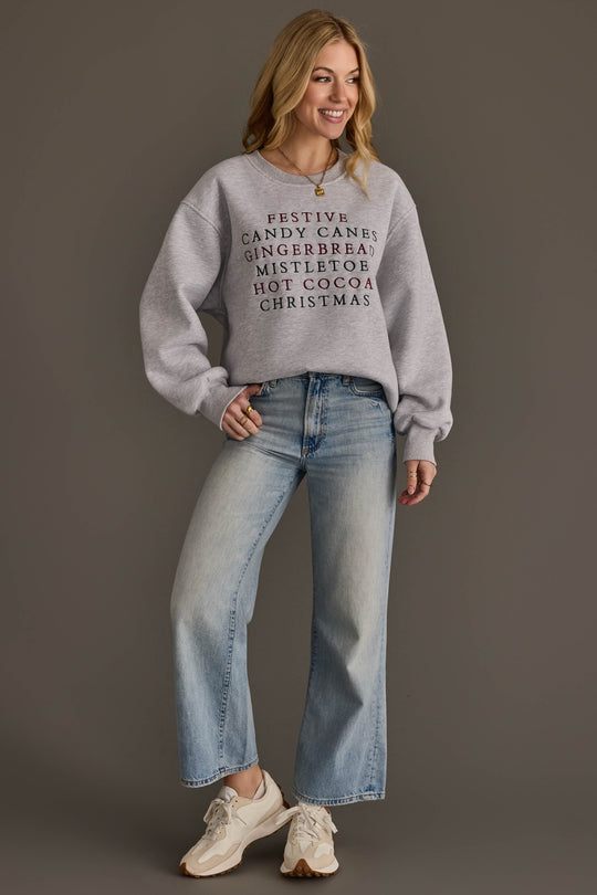 A woman wears a Christmas List Embroidered Sweatshirt featuring a list of embroidered words in red and black, paired with light-wash wide-leg jeans and athletic sneakers.