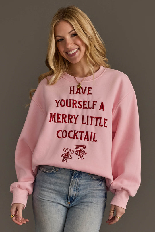 A woman wears a pink Pink Merry Little Cocktail Embroidered Sweatshirt featuring the red text HAVE YOURSELF A MERRY LITTLE COCKTAIL above an illustration of two martini glasses.