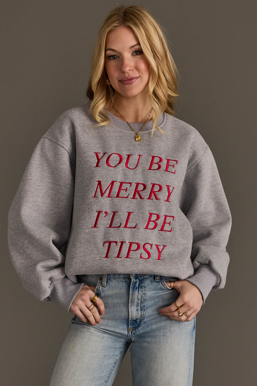 A person wearing the You Be Merry Embroidered Sweatshirt, a cozy, embroidered grey crewneck with the red text YOU BE MERRY ILL BE TIPSY.
