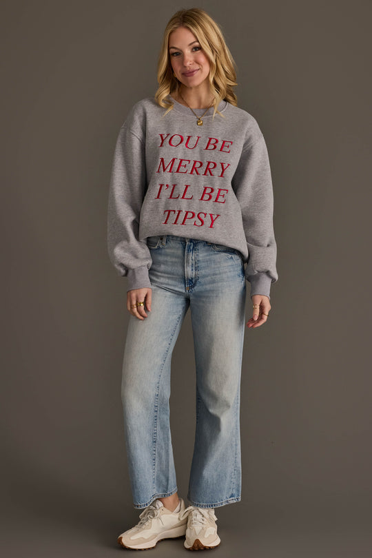 A woman wears a You Be Merry Embroidered Sweatshirt featuring the red text YOU BE MERRY ILL BE TIPSY, paired with light-wash jeans and athletic sneakers.