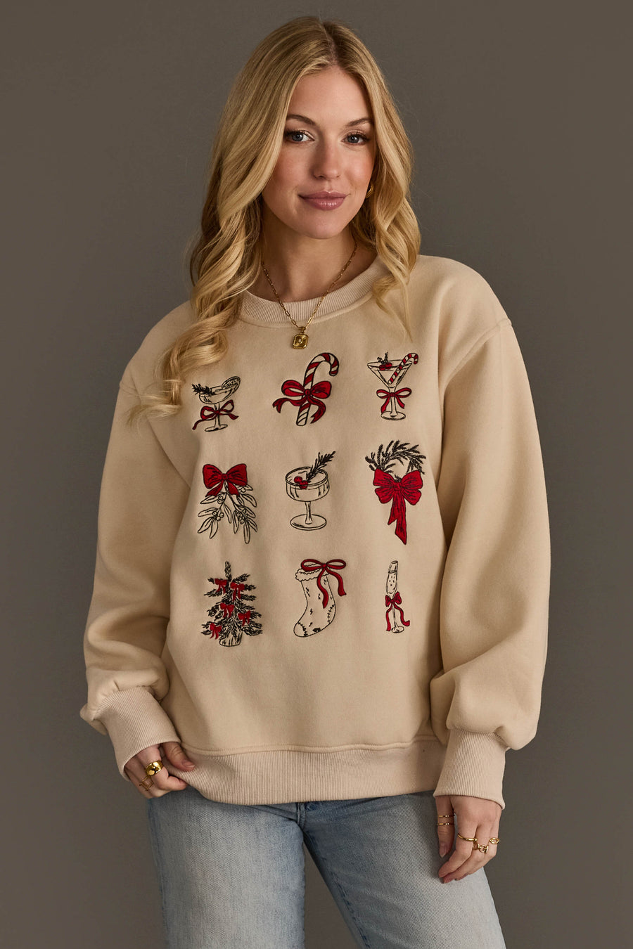 A woman wears a beige Christmas Graphics Embroidered Sweatshirt featuring red and green festive icons, including a candy cane, mistletoe, a stocking, and drinks.