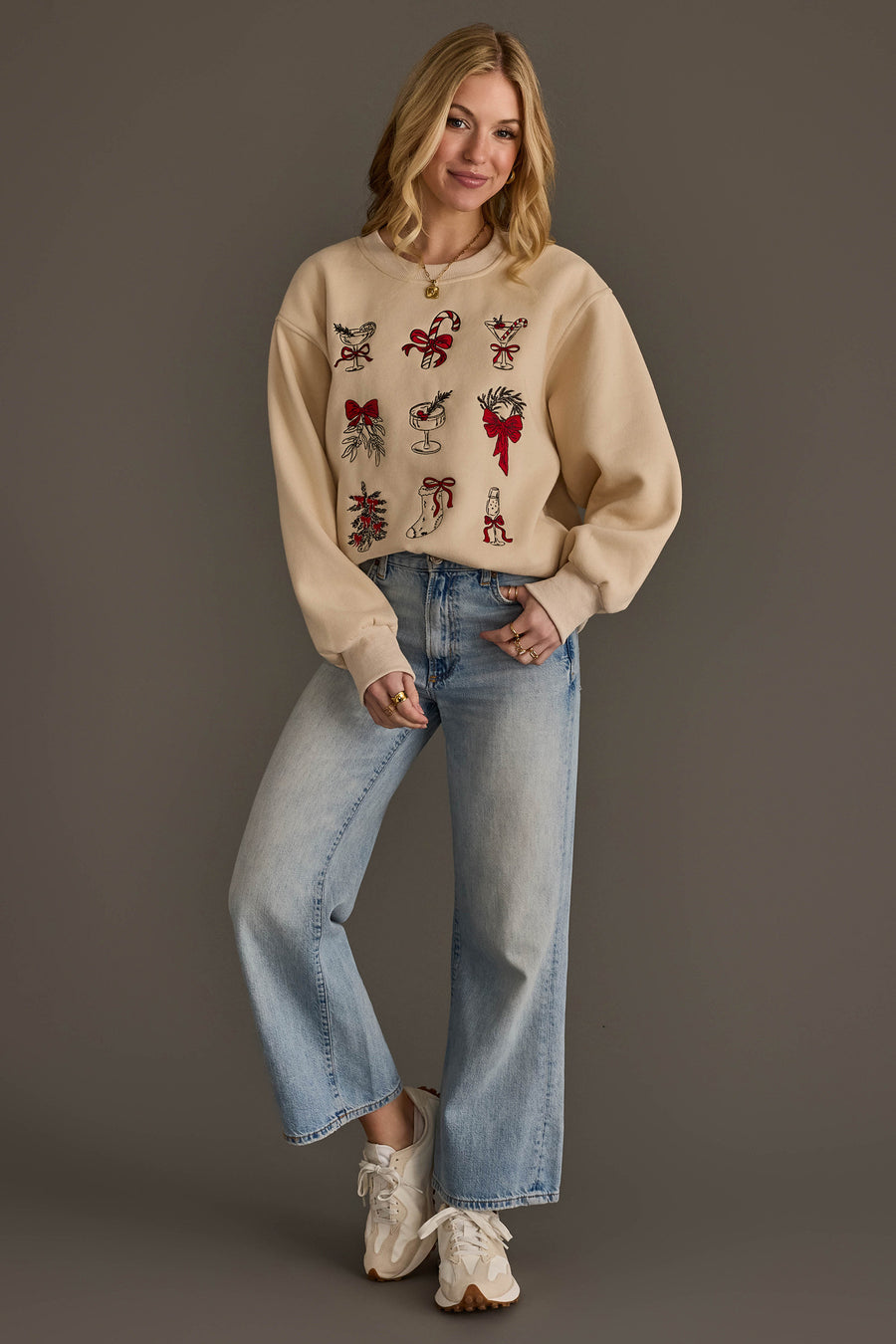 A woman wears a tan Christmas Graphics Embroidered Sweatshirt featuring red and green festive icons, paired with light-wash wide-leg jeans and sneakers.