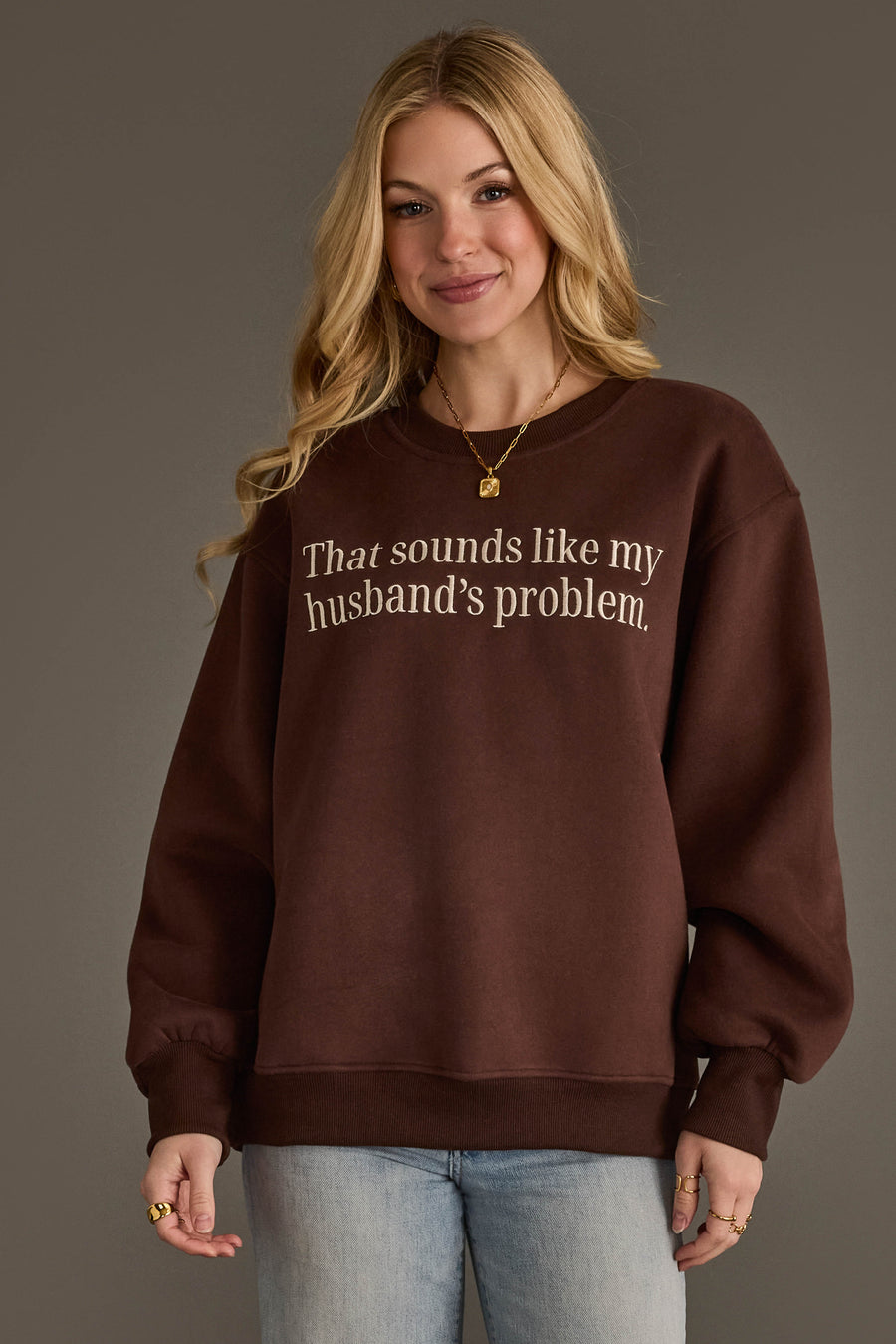 A woman with long blonde hair is smiling and standing against a plain background, wearing the Panache Apparel Collection’s Husband’s Problem Embroidered Sweatshirt in brown.