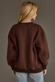 A person with long blonde hair is seen from behind, standing against a plain grey background while wearing the Panache Apparel Collection’s “Husband’s Problem Embroidered Sweatshirt” in brown, paired with light blue jeans.