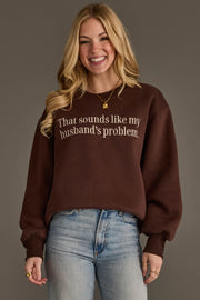 A smiling woman stands against a plain background, wearing light blue jeans and the Panache Apparel Collection’s “Husband’s Problem Embroidered Sweatshirt,” a brown sweatshirt featuring the phrase “That sounds like my husband’s problem.”.