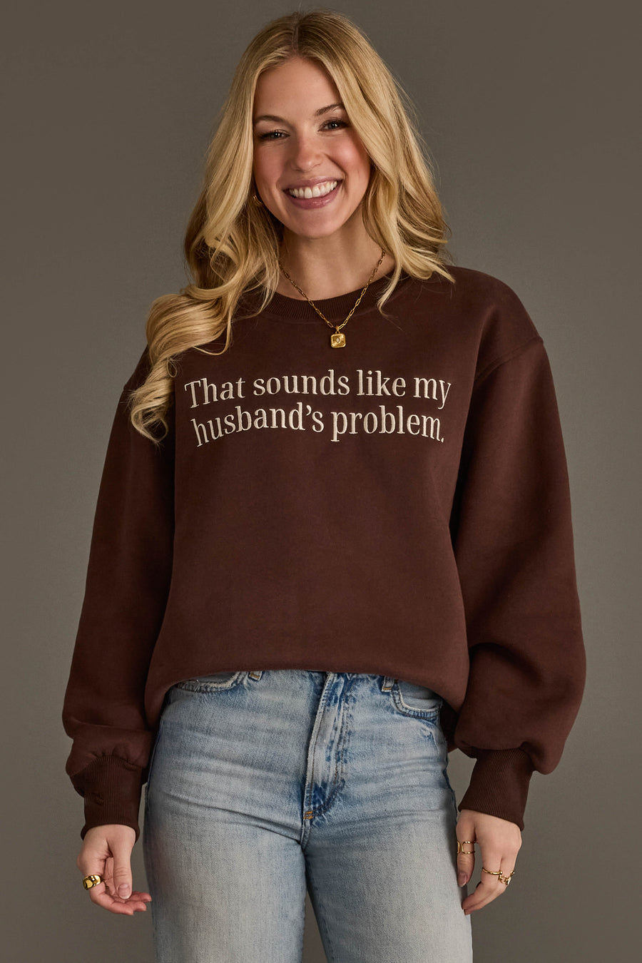A smiling woman stands against a plain background, wearing light blue jeans and the Panache Apparel Collection’s “Husband’s Problem Embroidered Sweatshirt,” a brown sweatshirt featuring the phrase “That sounds like my husband’s problem.”.