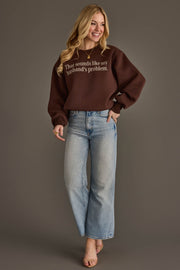 A smiling woman with long blonde hair wears the Panache Apparel Collection’s Husband’s Problem Embroidered Sweatshirt in brown, paired with light blue jeans and heels, standing against a plain gray backdrop.