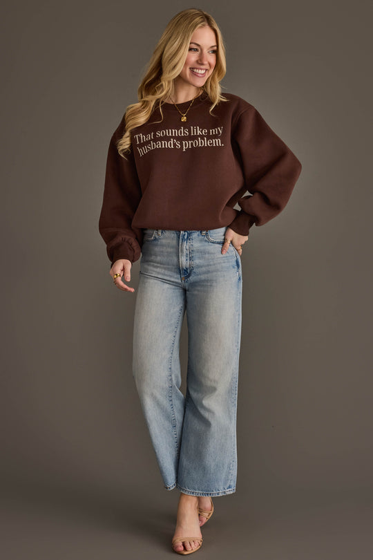 A smiling woman with long blonde hair wears the Panache Apparel Collection’s Husband’s Problem Embroidered Sweatshirt in brown, paired with light blue jeans and heels, standing against a plain gray backdrop.