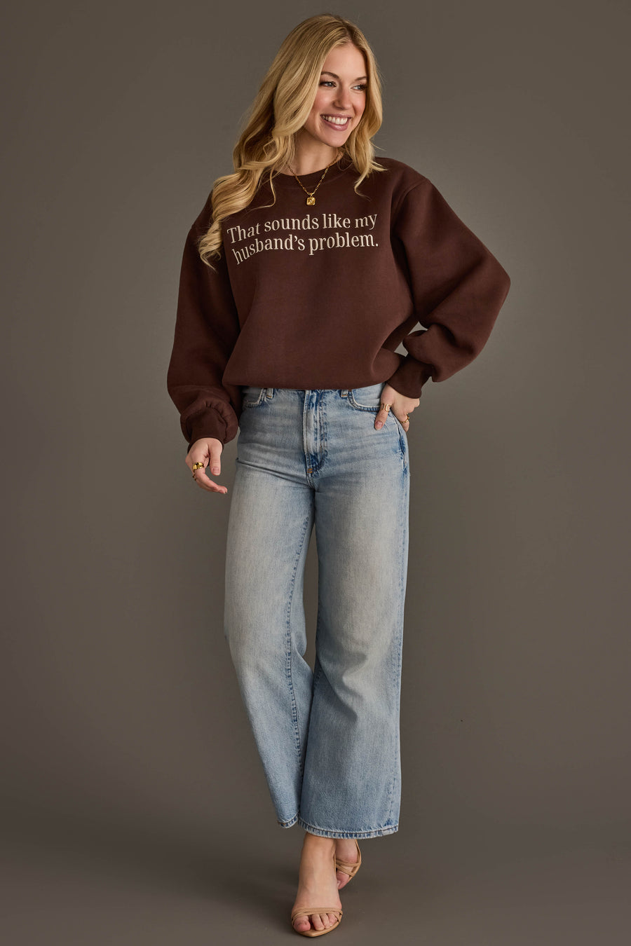 A smiling woman with long blonde hair wears the Panache Apparel Collection’s Husband’s Problem Embroidered Sweatshirt in brown, paired with light blue jeans and heels, standing against a plain gray backdrop.