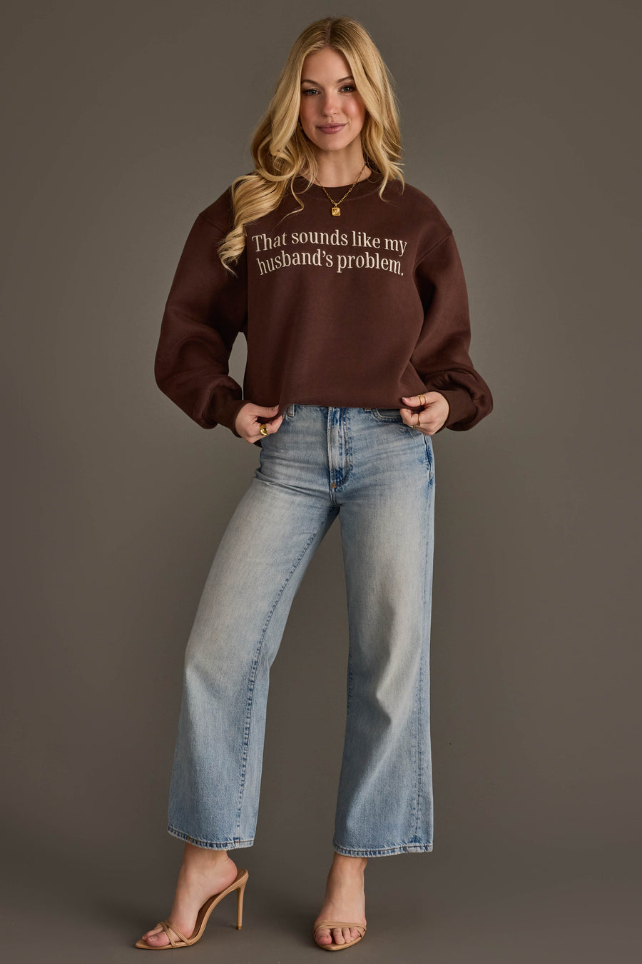 A woman with long blonde hair is smiling as she stands against a plain background, wearing the Husband’s Problem Embroidered Sweatshirt by Panache Apparel Collection in brown, paired with light blue jeans and beige heeled sandals.
