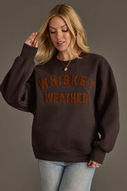 A woman with blonde hair wears the Panache Apparel Collection Charcoal & Camel Whiskey Weather Sweatshirt, featuring textured orange lettering. She pairs it with blue jeans and gold necklaces while standing against a plain gray background.