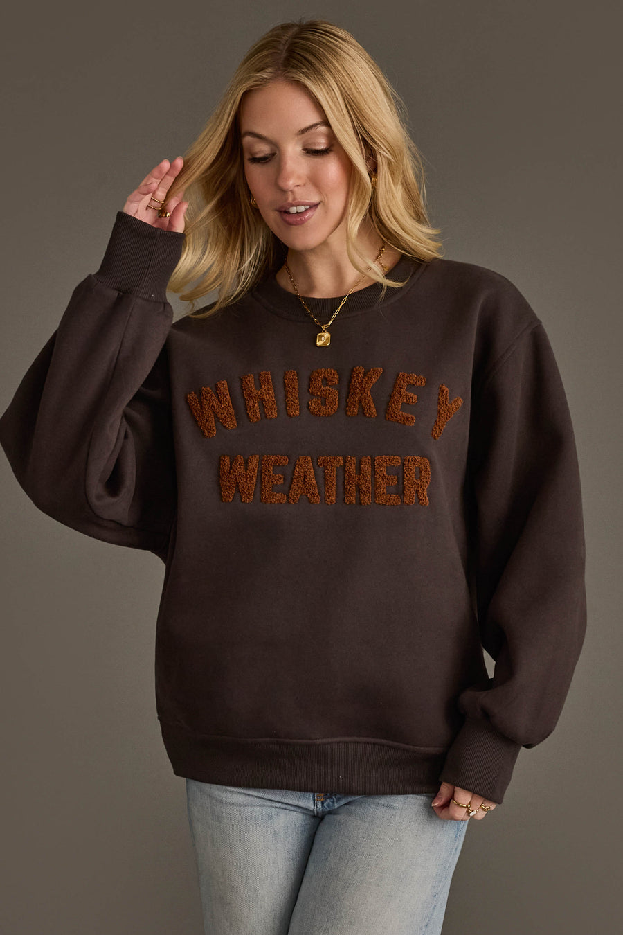 A woman with blonde hair wears the Panache Apparel Collection Charcoal & Camel Whiskey Weather Sweatshirt, featuring textured orange lettering. She pairs it with blue jeans and gold necklaces while standing against a plain gray background.