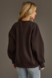 A person with long blonde hair wears the Panache Apparel Collection’s Charcoal & Camel Whiskey Weather Sweatshirt with light blue jeans, standing against a plain background and facing away from the camera.