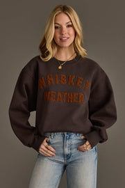 A woman smiles at the camera while wearing the Panache Apparel Collection’s Charcoal & Camel Whiskey Weather Sweatshirt, styled with light blue jeans and gold jewelry, against a plain gray background.