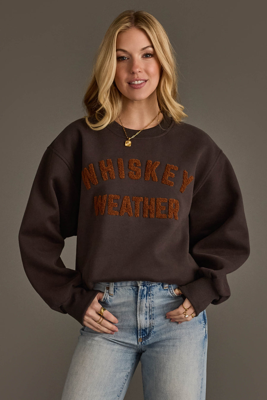 A woman smiles at the camera while wearing the Panache Apparel Collection’s Charcoal & Camel Whiskey Weather Sweatshirt, styled with light blue jeans and gold jewelry, against a plain gray background.