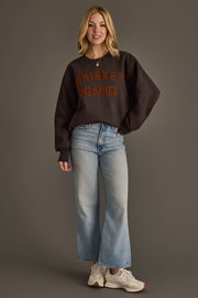 A woman stands against a grey background, wearing the Panache Apparel Collection’s Charcoal & Camel Whiskey Weather Sweatshirt, paired with light blue jeans and white sneakers. She smiles, her left hand in her pocket and blonde hair loose around her shoulders.
