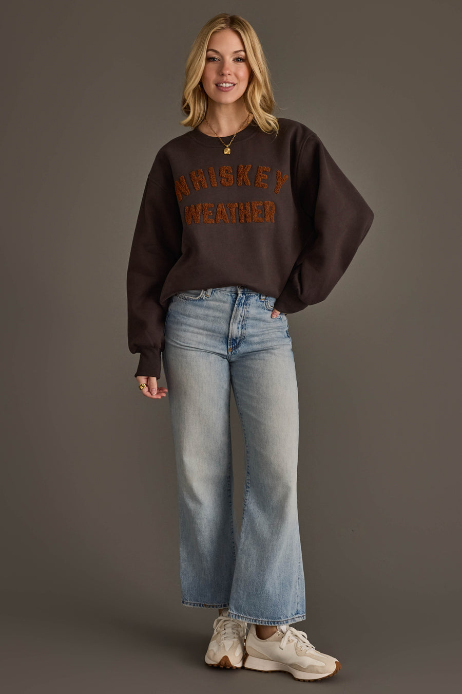 A woman stands against a grey background, wearing the Panache Apparel Collection’s Charcoal & Camel Whiskey Weather Sweatshirt, paired with light blue jeans and white sneakers. She smiles, her left hand in her pocket and blonde hair loose around her shoulders.