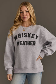 A woman with long blonde hair stands against a neutral background, smiling with one hand on her hip while wearing the Panache Apparel Collection Light Gray & Dark Gray Whiskey Weather Sweatshirt featuring bold black lettering.