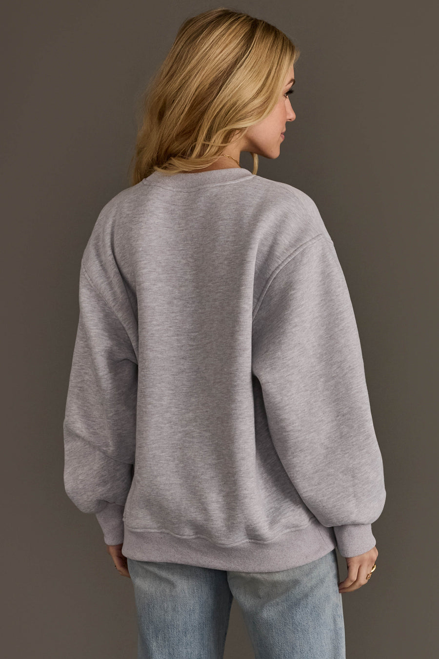 Wearing the Panache Apparel Collection’s Light Gray & Dark Gray Whiskey Weather Sweatshirt, a person with long blonde hair and light blue jeans stands facing away from the camera against a plain gray background.