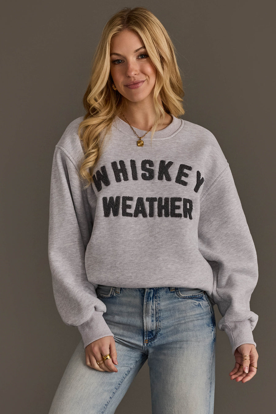 A smiling woman with long blonde hair is wearing the Panache Apparel Collection’s Light Gray & Dark Gray Whiskey Weather Sweatshirt, featuring ‘WHISKEY WEATHER’ in bold black letters, paired with light blue jeans and standing against a plain brown background.