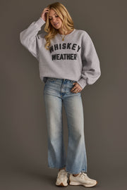 A woman wearing the Panache Apparel Collection Light Gray & Dark Gray Whiskey Weather Sweatshirt, paired with light blue wide-leg jeans and white sneakers, stands against a plain brown background, touching her hair and smiling slightly.