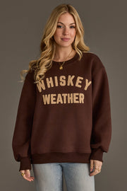 A woman with long blonde hair stands against a plain background, wearing the Panache Apparel Collection’s Dark Brown & Tan Whiskey Weather Sweatshirt featuring large tan lettering, paired with light blue jeans and gold jewelry.