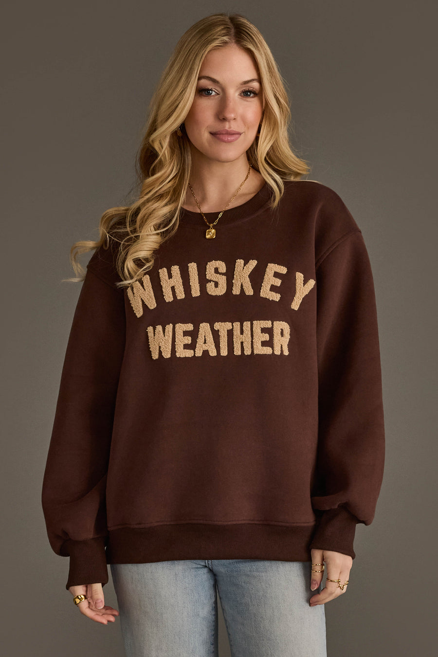 A woman with long blonde hair stands against a plain background, wearing the Panache Apparel Collection’s Dark Brown & Tan Whiskey Weather Sweatshirt featuring large tan lettering, paired with light blue jeans and gold jewelry.