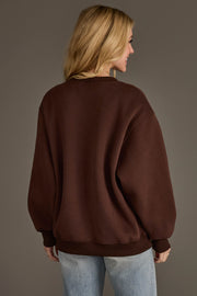 A woman with long blonde hair stands with her back to the camera against a plain background, wearing the Panache Apparel Collection’s Dark Brown & Tan Whiskey Weather Sweatshirt paired with light blue jeans.