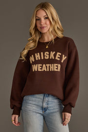 A woman with long blonde hair wears the Panache Apparel Collection’s Dark Brown & Tan Whiskey Weather Sweatshirt paired with light blue jeans, standing against a plain gray background.