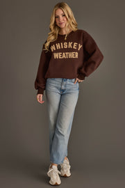 A woman stands against a gray background wearing a Panache Apparel Collection Dark Brown & Tan Whiskey Weather Sweatshirt, light blue jeans, and white sneakers. She has long blonde hair and is smiling slightly.