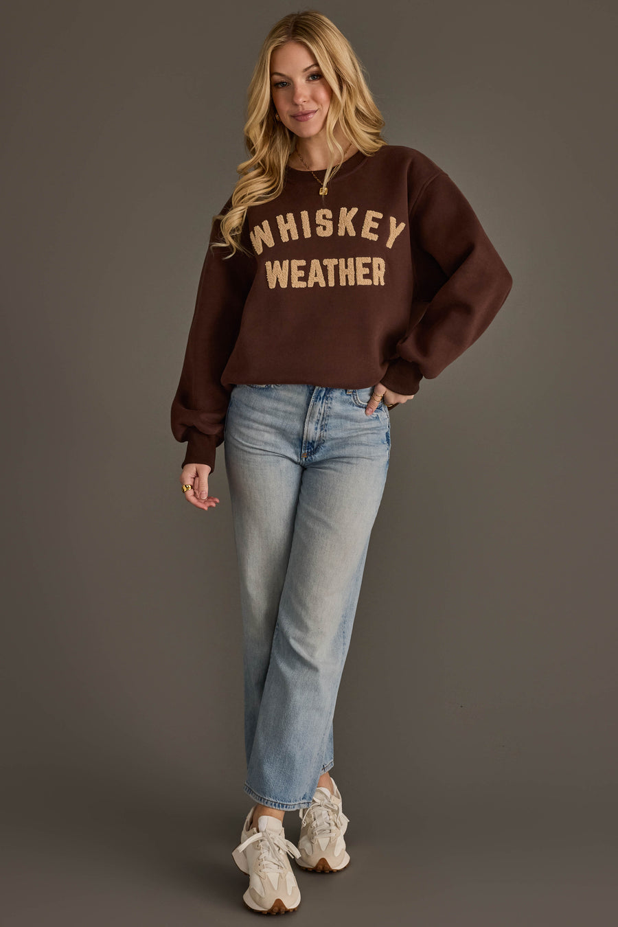 A woman stands against a gray background wearing a Panache Apparel Collection Dark Brown & Tan Whiskey Weather Sweatshirt, light blue jeans, and white sneakers. She has long blonde hair and is smiling slightly.