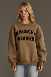 A woman with blonde hair is wearing the Panache Apparel Collection’s Taupe & Dark Brown Whiskey Weather Sweatshirt, featuring bold black lettering. She pairs it with light blue jeans and gold jewelry, standing against a plain brown background.