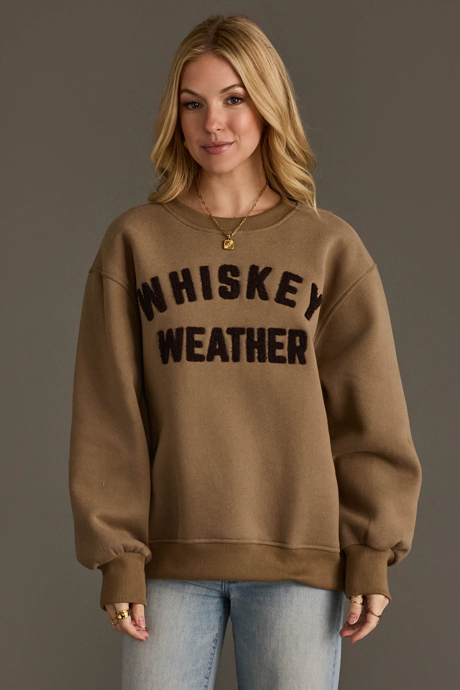 A woman with blonde hair is wearing the Panache Apparel Collection’s Taupe & Dark Brown Whiskey Weather Sweatshirt, featuring bold black lettering. She pairs it with light blue jeans and gold jewelry, standing against a plain brown background.