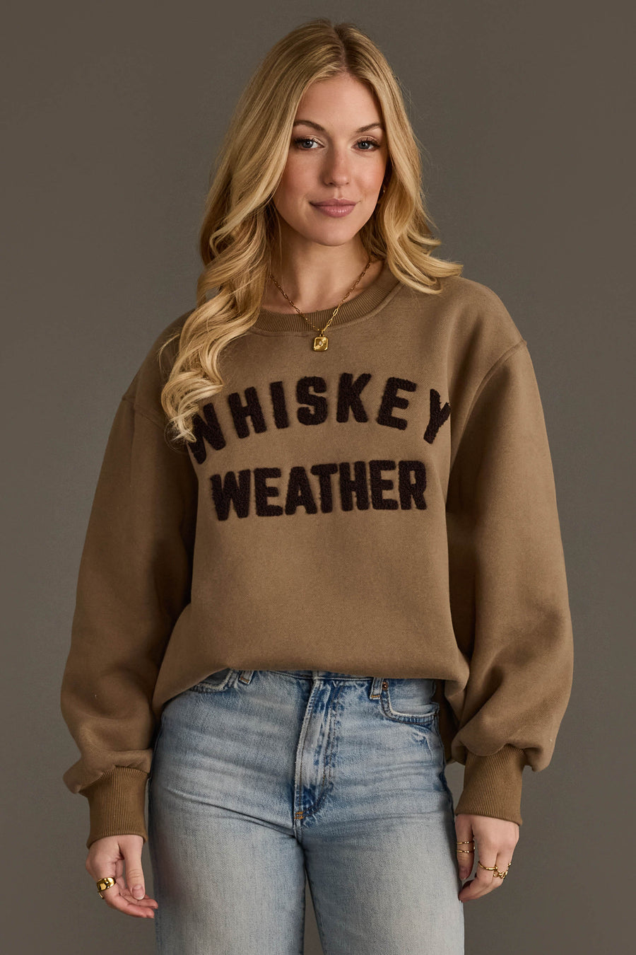 A woman with long blonde hair wears the Panache Apparel Collection’s Taupe & Dark Brown Whiskey Weather Sweatshirt, paired with light blue jeans, standing against a plain gray background.