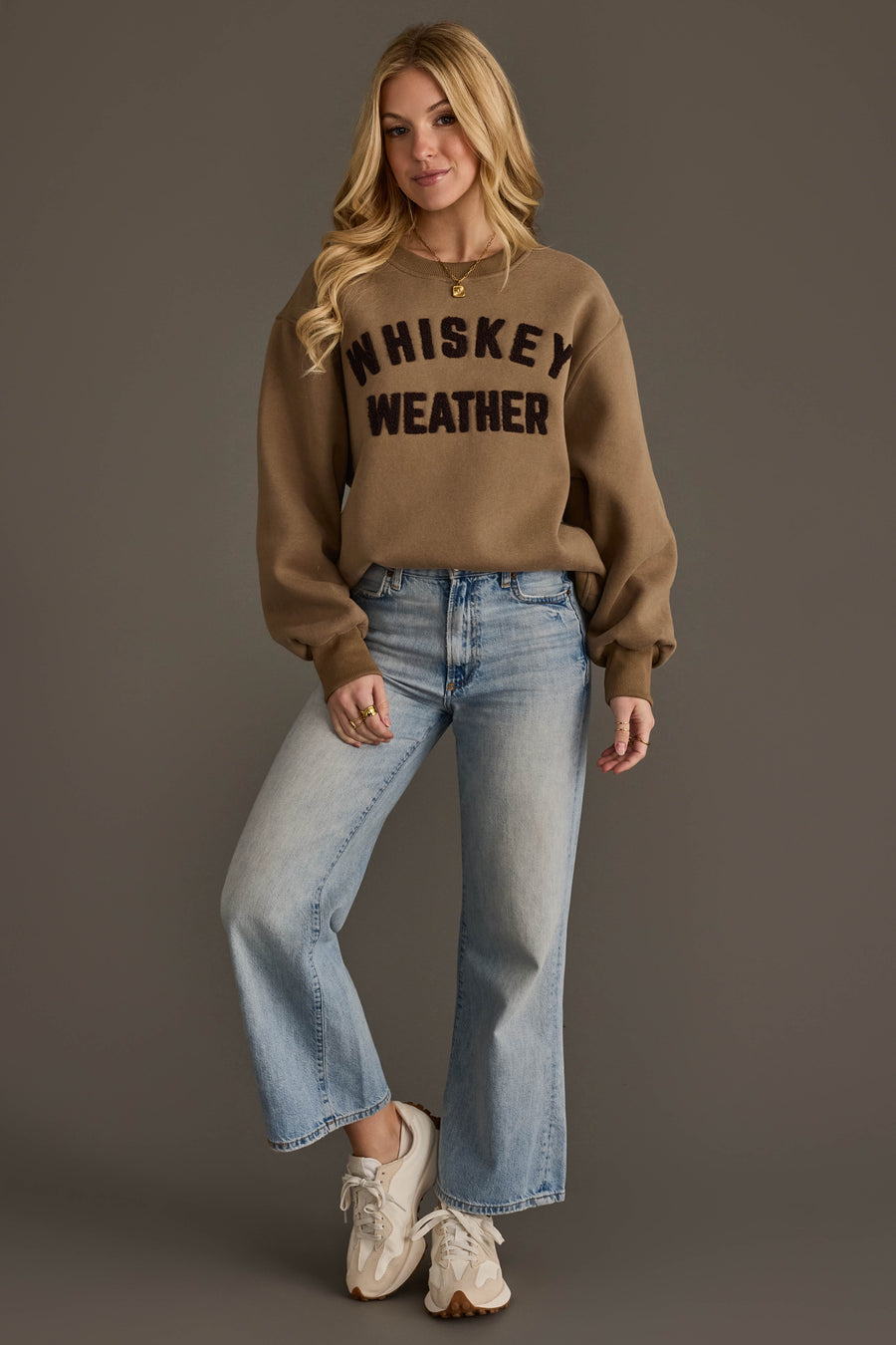 A woman stands against a plain background, wearing the Panache Apparel Collection’s Taupe & Dark Brown Whiskey Weather Sweatshirt, paired with light blue jeans and white sneakers. She has long blonde hair and a relaxed pose.