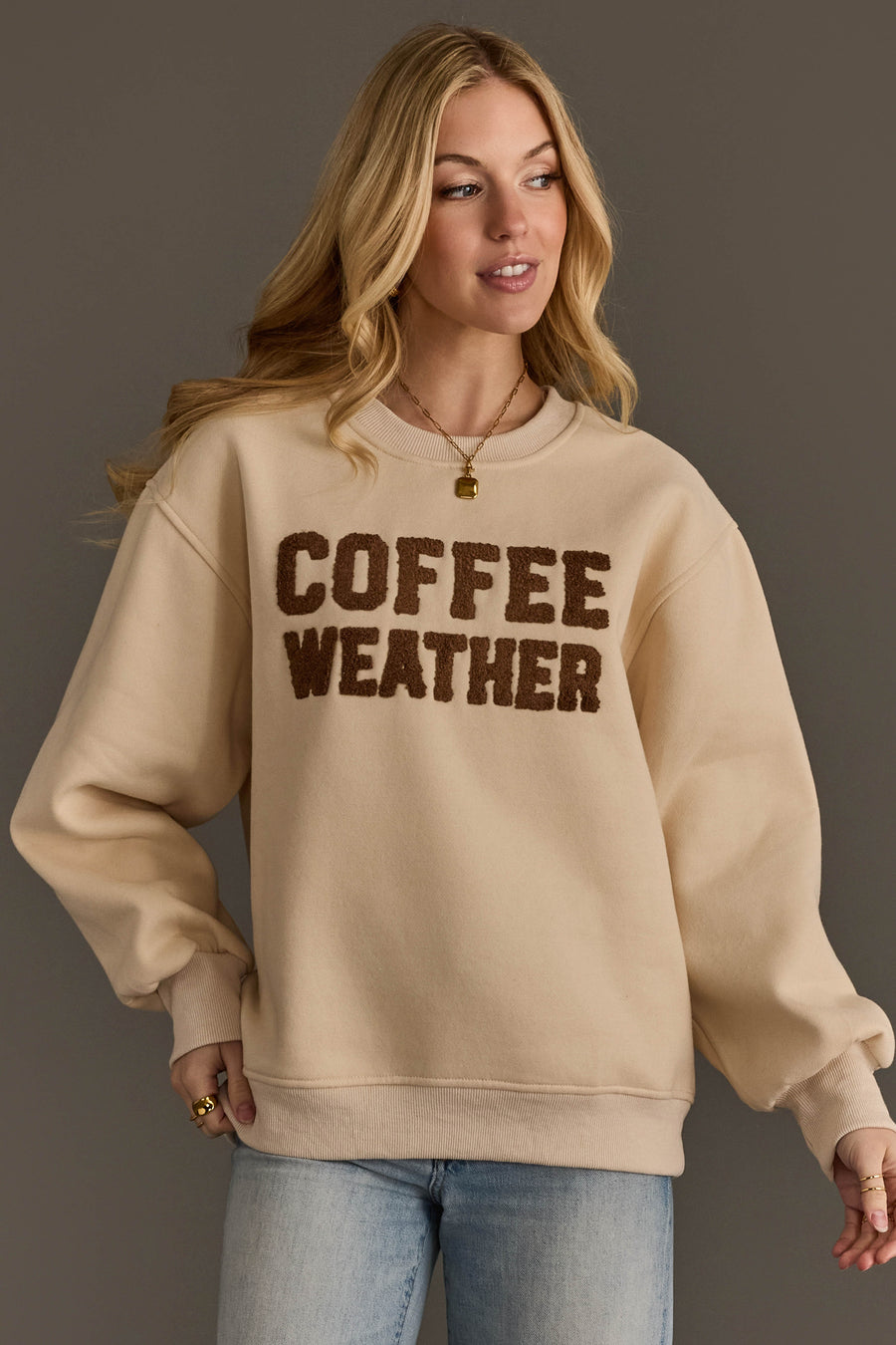 A woman stands against a dark background wearing the Panache Apparel Collection’s Beige & Dark Brown Coffee Weather Sweatshirt, styled with light blue jeans and gold jewelry.