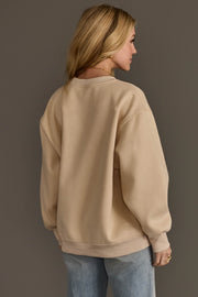 A woman with long blonde hair, wearing the Panache Apparel Collection’s Beige & Dark Brown Coffee Weather Sweatshirt paired with light blue jeans, stands facing away from the camera against a plain brown background.