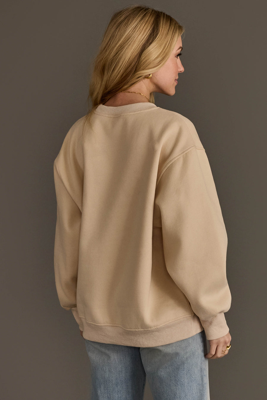 A woman with long blonde hair, wearing the Panache Apparel Collection’s Beige & Dark Brown Coffee Weather Sweatshirt paired with light blue jeans, stands facing away from the camera against a plain brown background.