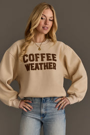 A woman wearing the Panache Apparel Collection’s Beige & Dark Brown Coffee Weather Sweatshirt, paired with blue jeans and gold jewelry, stands against a neutral background.