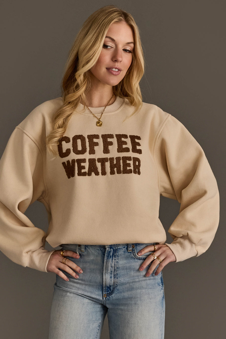 A woman wearing the Panache Apparel Collection’s Beige & Dark Brown Coffee Weather Sweatshirt, paired with blue jeans and gold jewelry, stands against a neutral background.