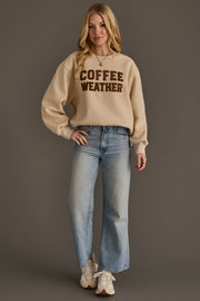 A woman poses against a brown backdrop, dressed in the Beige & Dark Brown Coffee Weather Sweatshirt from Panache Apparel Collection, paired with light blue jeans and white sneakers. She has long blonde hair and a soft smile.