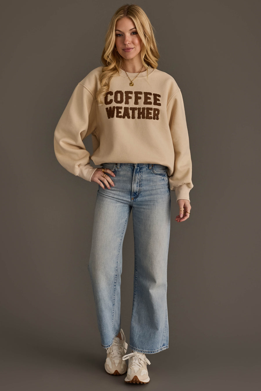 A woman poses against a brown backdrop, dressed in the Beige & Dark Brown Coffee Weather Sweatshirt from Panache Apparel Collection, paired with light blue jeans and white sneakers. She has long blonde hair and a soft smile.