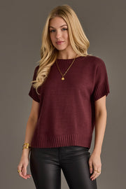 A woman with long blonde hair wears the Panache Apparel Collection Maroon Short Sleeve Sweater, paired with black leather pants and gold jewelry, standing against a plain gray background.