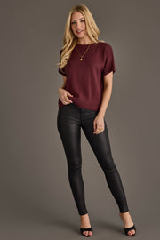 A woman with long blonde hair wears the Maroon Short Sleeve Sweater from Panache Apparel Collection, paired with black leather pants, black heels, and gold jewelry. She stands against a plain brown background, looking at the camera and smiling slightly.