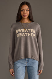 A woman stands facing forward against a plain gray background, wearing the Panache Apparel Collection’s Sweater Weather Sweater in gray, paired with light blue jeans and layered gold necklaces.