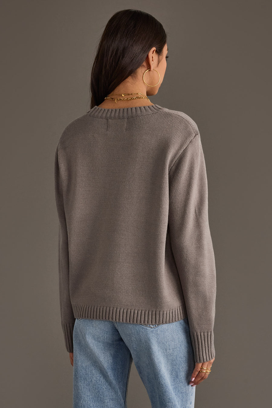 A woman with long brown hair, wearing large hoop earrings, models the Sweater Weather Sweater from Panache Apparel Collection in taupe, paired with light blue jeans. She stands with her back to the camera against a plain gray background.