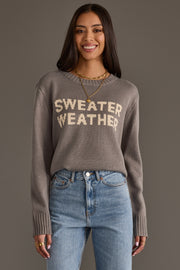 A woman stands and smiles against a plain gray background, wearing the Panache Apparel Collection "Sweater Weather" sweater in gray with beige lettering, paired with blue jeans.