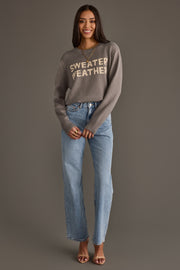A woman stands against a gray background, smiling with her hands clasped in front, wearing the Panache Apparel Collection Sweater Weather Sweater in gray, paired with light blue jeans and brown heeled sandals.