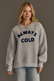 A woman with long blonde hair wears the Panache Apparel Collection’s Always Cold Sweatshirt—a grey pullover featuring “ALWAYS COLD” in large, dark blue letters—while standing against a plain brown background.